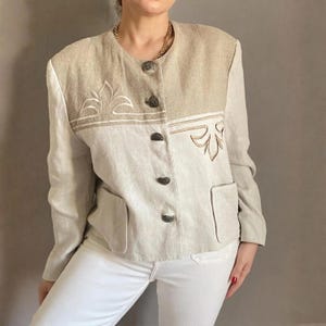 May include: Beige and tan blazer with embroidered floral details. The jacket features a two-tone design, with a lighter beige on the lower half and a darker tan on the upper half. It has four dark buttons and two front pockets. The blazer is a vintage style.
