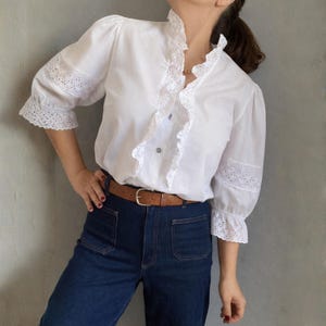 Op de afbeelding: Witte blouse met een gerimpelde kraag en mouwen, voorzien van eyelet-kant details. De blouse met driekwart mouwen wordt gecombineerd met een high-waisted, donkerblauwe denim jeans en een bruine leren riem.