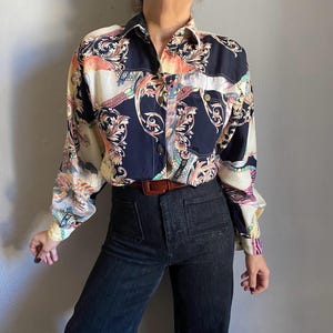 Pode incluir: Uma camisa abotoada com fundo azul marinho escuro e um padrão floral colorido. A camisa de manga comprida tem um cinto castanho e é combinada com jeans escuros. O design da camisa inclui tons de pêssego, creme e verde-azulado.