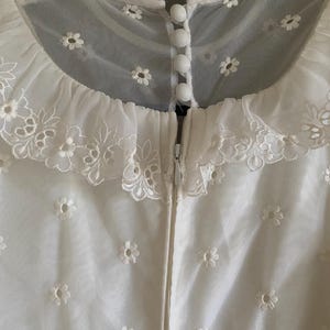 Op de afbeelding: Witte kantenjurk met een gegolfde kraag en een ritssluiting aan de achterkant. De jurk is bedekt met kleine witte geborduurde bloemen.