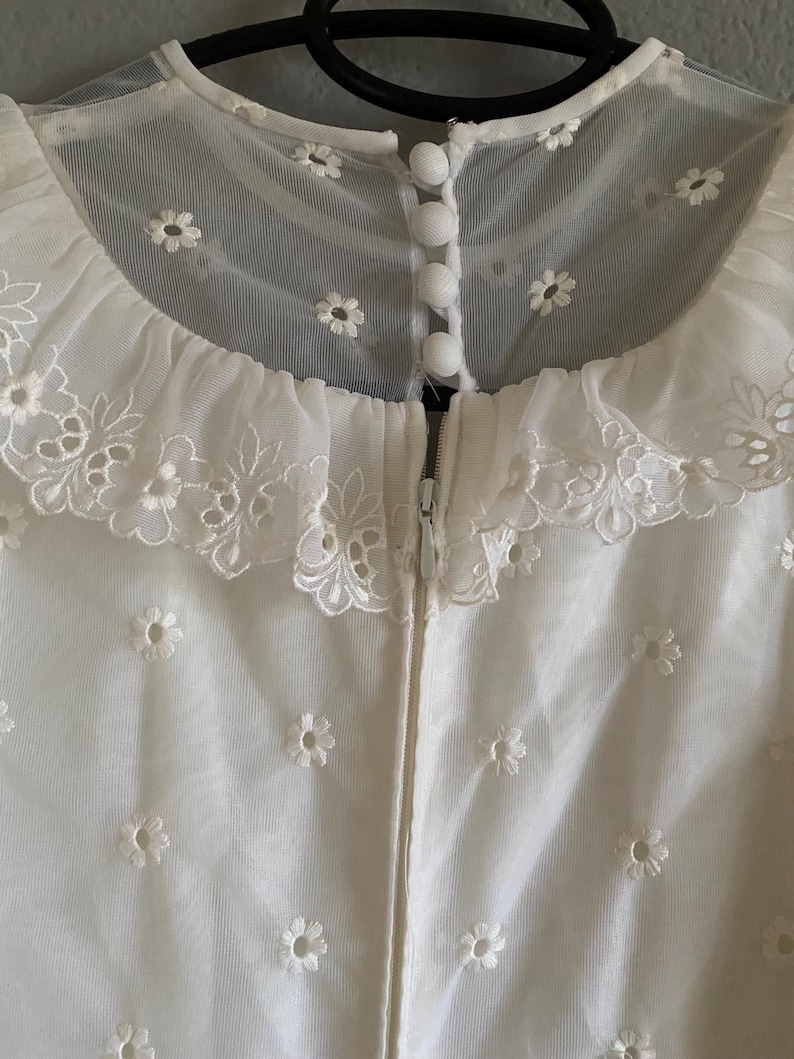 Op de afbeelding: Witte kantenjurk met een gegolfde kraag en een ritssluiting aan de achterkant. De jurk is bedekt met kleine witte geborduurde bloemen.