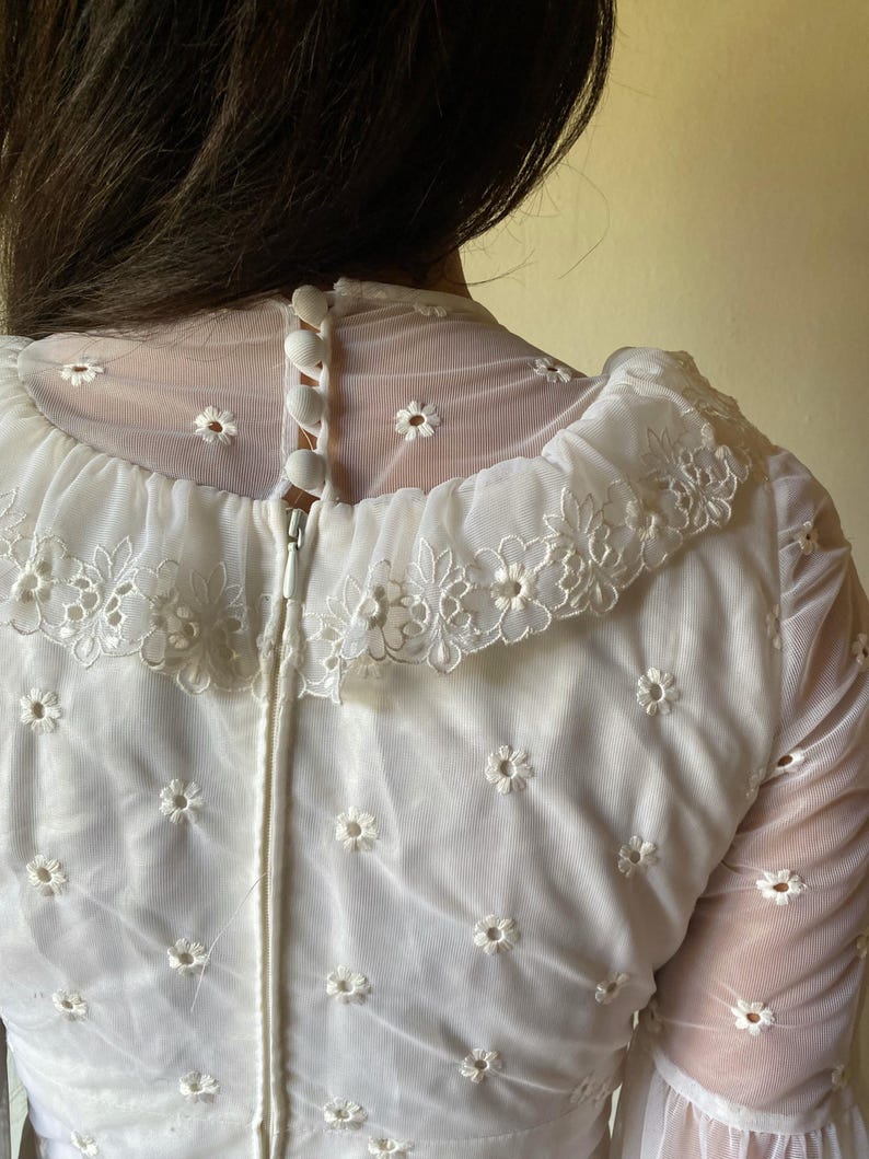 Op de afbeelding: Witte kantenjurk met een knoopsluiting aan de achterkant van de hals en een rits aan de achterkant. De jurk heeft een delicaat bloemenpatroon en een gerimpelde kraag.