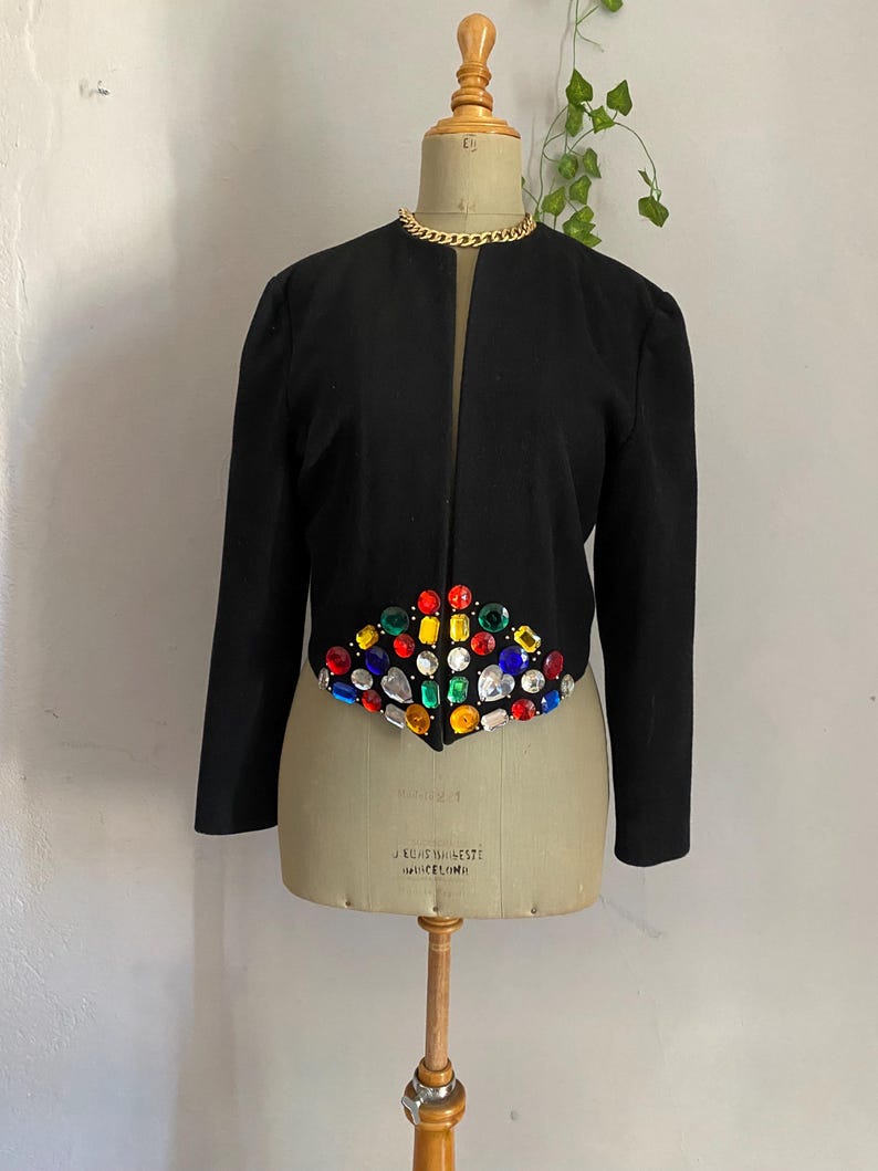 Puede incluir: Chaqueta negra con un dise&ntilde;o con incrustaciones de joyas. La chaqueta presenta un dise&ntilde;o sin cuello y mangas largas. Los adornos de joyas son de varios colores, incluyendo rojo, azul, verde y amarillo. Un collar de cadena dorada rodea el cuello del maniqu&iacute;.