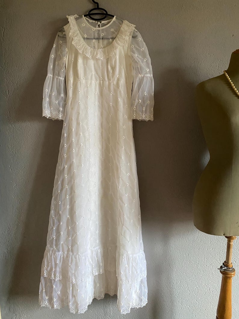 Op de afbeelding: Een witte, lange, geborduurde jurk met een gerimpelde halslijn en een gelaagde rok.