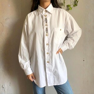 May include: White button-down shirt with long sleeves and a chest pocket. The shirt features embroidered floral detailing down the front and gold-coloured buttons. The shirt is worn with blue jeans.