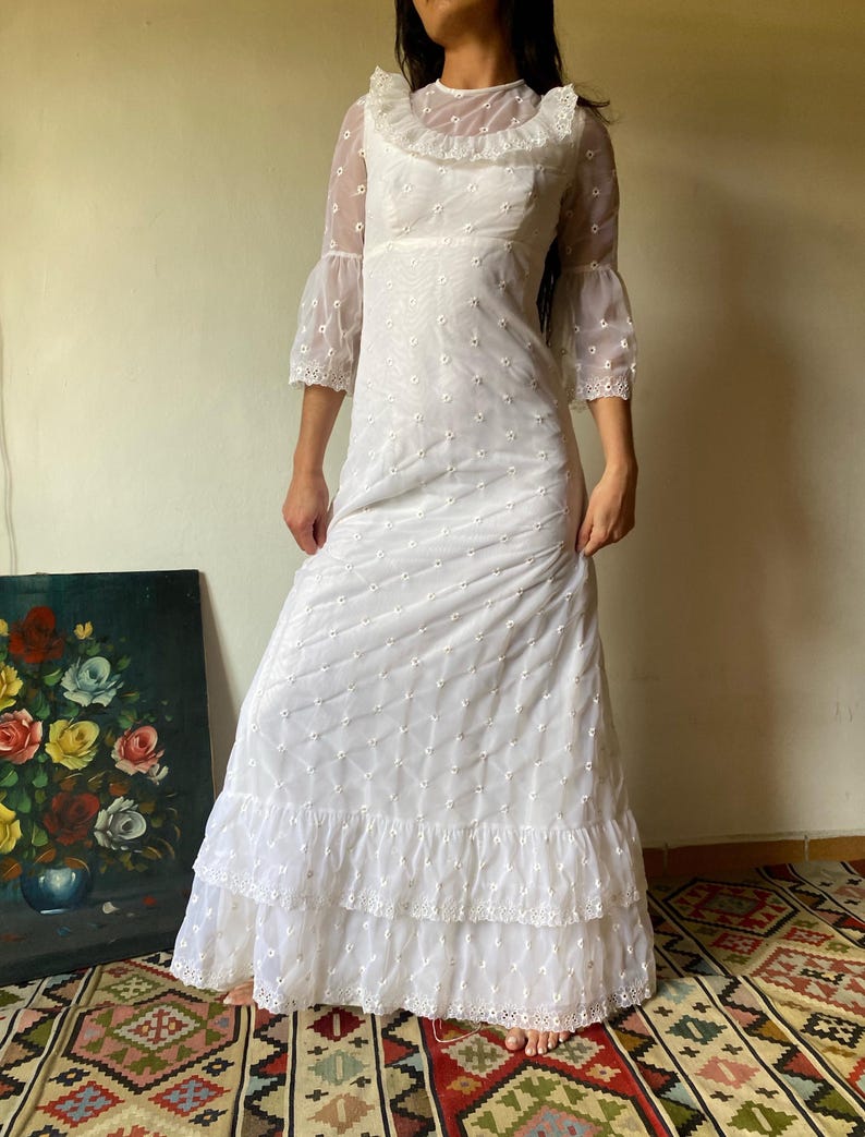 Op de afbeelding: Een witte maxi-jurk met een transparante overlay en kantversiering. De jurk heeft een ronde hals, 3/4 mouwen en een gelaagde rok.
