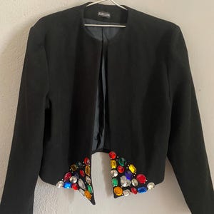 Puede incluir: Una chaqueta negra con un dise&ntilde;o adornado con joyas. La chaqueta presenta un dise&ntilde;o sin cuello y mangas largas. Los bordes delanteros inferiores est&aacute;n adornados con una variedad de gemas coloridas y facetadas de varias formas y tama&ntilde;os. La marca "BLOSGUIN" es visible.