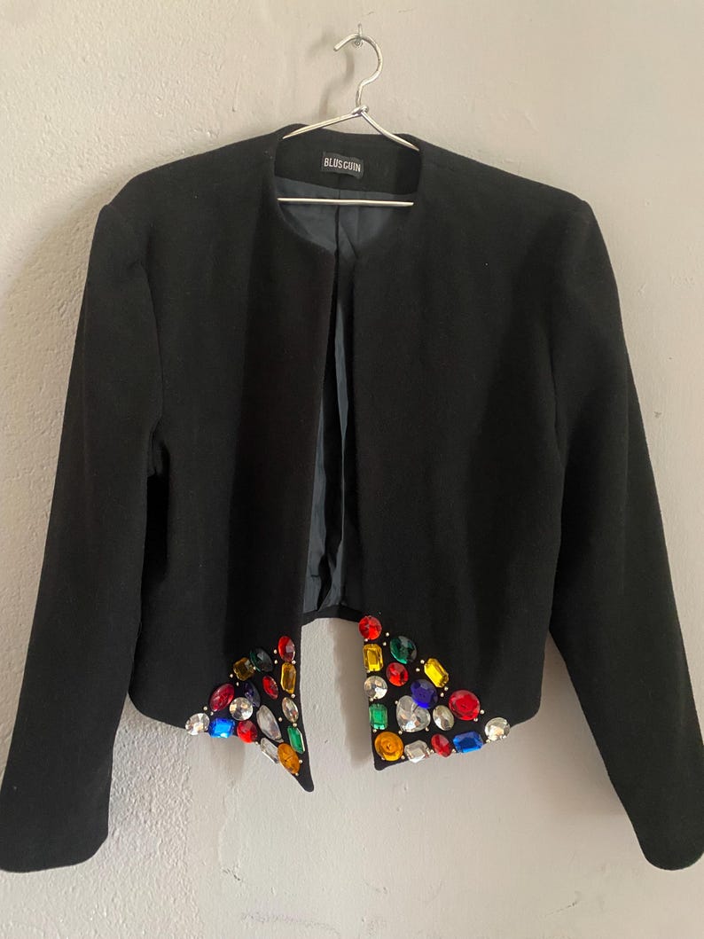 Puede incluir: Una chaqueta negra con un dise&ntilde;o adornado con joyas. La chaqueta presenta un dise&ntilde;o sin cuello y mangas largas. Los bordes delanteros inferiores est&aacute;n adornados con una variedad de gemas coloridas y facetadas de varias formas y tama&ntilde;os. La marca "BLOSGUIN" es visible.