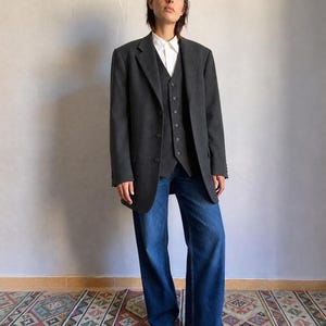 Peut inclure: Un blazer gris anthracite porté sur un gilet assorti boutonné et une chemise blanche à col rigide, associé à un jean bleu à jambes larges. L'ensemble comprend un blazer structuré à revers crantés et un gilet à cinq boutons.