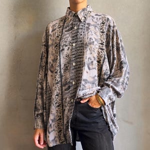 May include: A long-sleeved button-down shirt featuring a grey, black, and white abstract pattern with vertical panels. The shirt has a standard collar, a front button placket, and long sleeves with buttoned cuffs, paired with solid black trousers.