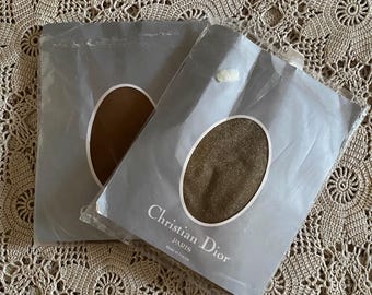 May include: Two pairs of Christian Dior pantyhose in their original packaging. The packaging is gray with a clear window showing the color of the hosiery. The text "Christian Dior PARIS Made in France" is printed on the packaging. The pantyhose appear to be a bronze color.
