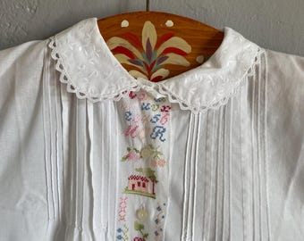 May include: White vintage blouse with a Peter Pan collar and vertical pleats. The blouse features a colorful cross-stitch design down the front with floral and house motifs. The blouse is hanging on a wooden hanger with a painted floral design.