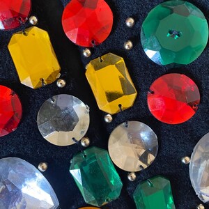 Puede incluir: Primer plano de una tela azul oscuro adornada con varias piedras preciosas de colores y cuentas plateadas. Las piedras preciosas son en tonos de rojo, amarillo, verde y plata, con diferentes formas y tama&ntilde;os. Las cuentas son peque&ntilde;as y redondas.