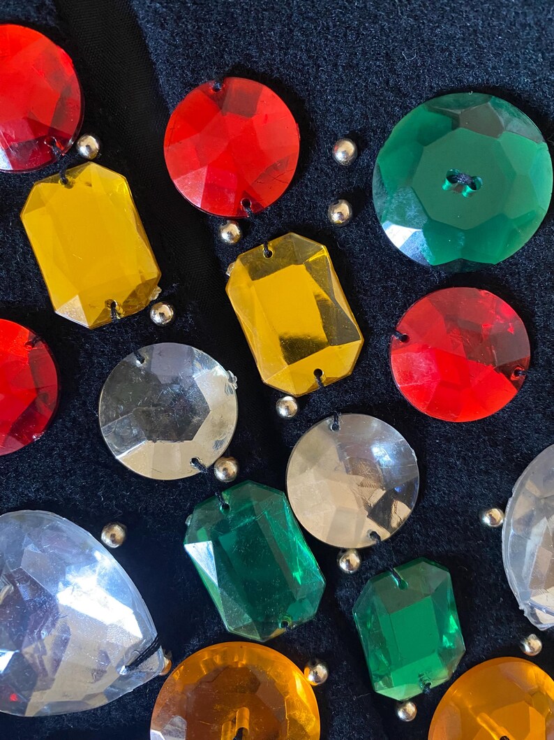 Puede incluir: Primer plano de una tela azul oscuro adornada con varias piedras preciosas de colores y cuentas plateadas. Las piedras preciosas son en tonos de rojo, amarillo, verde y plata, con diferentes formas y tama&ntilde;os. Las cuentas son peque&ntilde;as y redondas.