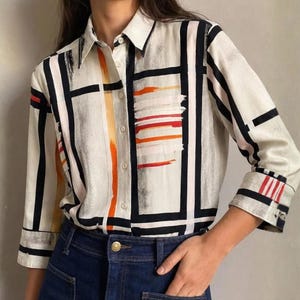 May include: A long-sleeved button-down shirt in an off-white colour featuring a bold black grid pattern. The design includes horizontal brushstroke accents in vibrant orange and red across the chest and cuffs, with a classic collar and buttons.