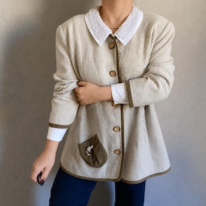 May include: Beige blazer with a white lace collar, brown trim, and wooden buttons. The jacket has a small pocket with a decorative element. The sleeves are long, and the blazer is worn over a white shirt.