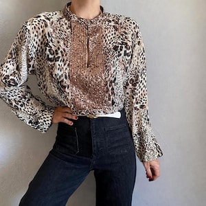 Op de afbeelding: Een blouse met lange mouwen en een luipaardprint in bruin, beige en zwart. De blouse heeft een hoge kraag en een paneel met pailletten aan de voorkant. Het model draagt een donkerblauwe jeans.