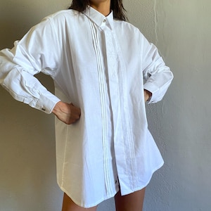 May include: A white long-sleeve shirt with a classic collar and buttoned cuffs. The shirt features vertical pleats down the front and delicate embroidered detailing. The shirt is made of a lightweight fabric.