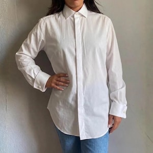 May include: A crisp white long-sleeved button-down shirt featuring a structured collar, a concealed front placket, and wide cuffs with single buttons. The shirt is crafted from a smooth, opaque fabric and is paired with blue denim jeans.