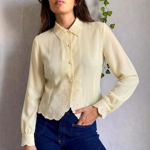 Op de afbeelding: Een lichtgele blouse met lange mouwen en een knoopsluiting. De blouse heeft een delicate, geborduurde kraag en zoom, met bijpassende details op de manchetten. Het model draagt de blouse met een donkerblauwe jeans.