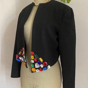 Puede incluir: Chaqueta bolero negra con mangas largas y cuello redondo. La chaqueta est&aacute; adornada con pedrer&iacute;a de colores joya a lo largo de los bordes delanteros. Se ve un collar de cadena dorada en el escote. La chaqueta se exhibe en un maniqu&iacute; vintage.