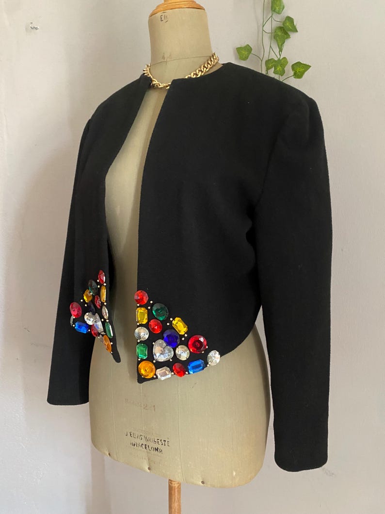 Puede incluir: Chaqueta bolero negra con mangas largas y cuello redondo. La chaqueta est&aacute; adornada con pedrer&iacute;a de colores joya a lo largo de los bordes delanteros. Se ve un collar de cadena dorada en el escote. La chaqueta se exhibe en un maniqu&iacute; vintage.