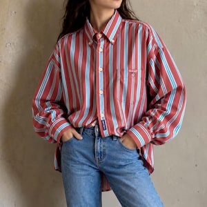 May include: A long-sleeve button-down shirt featuring vertical stripes in shades of red, light blue, and white. The shirt has a pointed collar, a single chest pocket with the word GANT embroidered in white, and blue denim jeans.