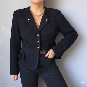 Könnte beinhalten: Ein dunkelblauer Blazer mit strukturiertem Stoff, rundem Kragen, dekorativen Knöpfen und Taschen Details. Der Blazer wird mit dunklen Jeans und einem grün-rot gestreiften Gürtel kombiniert.