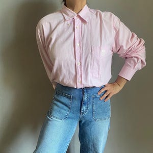 May include: A light pink button-down shirt with a pocket on the chest. The shirt has long sleeves and a button-down collar. The shirt is paired with blue denim jeans with large front pockets.