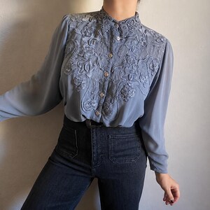 May include: A dusty blue long-sleeve button-down shirt featuring an intricate lace collar and a decorative embroidered floral panel across the chest. The garment has sheer sleeves and a button-up front with small, round, light-colored buttons.