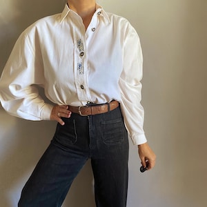 May include: White button-down shirt with long sleeves and decorative embroidery near the buttons. The shirt is paired with black denim jeans and a brown leather belt. The outfit is completed with a small black object.