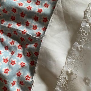 Op de afbeelding: Een stuk blauwe stof met een rood bloemenpatroon is genaaid aan een witte stof met een witte kantversiering.