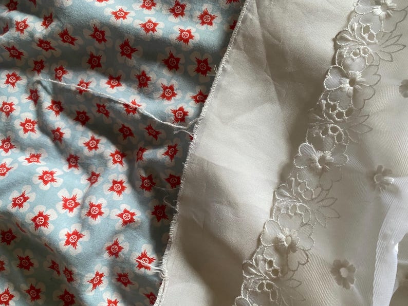 Op de afbeelding: Een stuk blauwe stof met een rood bloemenpatroon is genaaid aan een witte stof met een witte kantversiering.