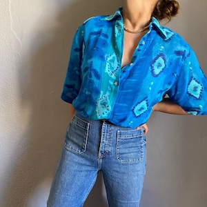 May include: A short-sleeved, button-down shirt in shades of blue with a patterned design. The shirt is paired with light-wash denim jeans. A gold-colored necklace is visible at the neckline. The outfit is casual and stylish.