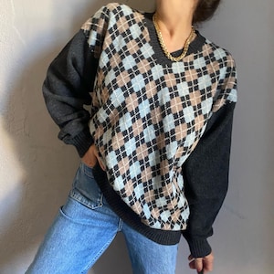 Könnte beinhalten: Ein Pullover mit Argyle-Muster in Blau-, Braun- und Schwarztönen. Die Ärmel sind dunkelgrau, und der Pullover wird mit einer blauen Jeans kombiniert. Eine goldene Halskette ist sichtbar.