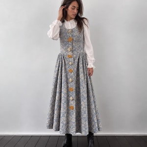 May include: A sleeveless floor-length dress in a blue and white floral pattern featuring a front button placket with alternating round mustard yellow and metallic silver buttons, worn over a white long-sleeved blouse with ruffled cuffs.