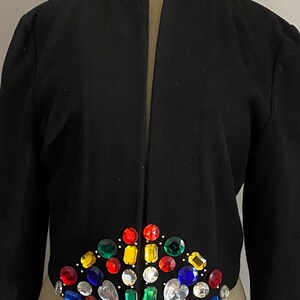 Puede incluir: Chaqueta negra con cuello de cadena dorada y adornos de piedras preciosas de colores. La chaqueta se exhibe en un maniqu&iacute;. El dise&ntilde;o de piedras preciosas presenta una variedad de formas y colores, incluyendo rojo, amarillo, verde y azul.