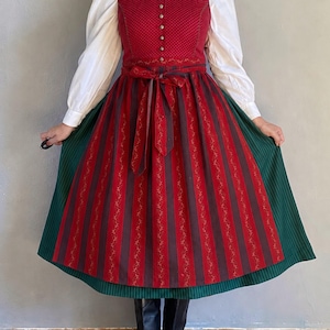 Op de afbeelding: Een jurk bestaande uit een witte blouse met lange mouwen, een rood lijfje en een groene rok. Het rode lijfje heeft een stippenpatroon en een rij metalen knopen. Een rood en donkerbruin gestreept schort met bloemmotief is in de taille gestrikt.