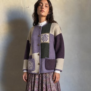 Könnte beinhalten: Eine langärmelige Strickjacke mit Knopfleiste und Patchwork-Design in den Farben Lavendel, Dunkelgrün, Beige und Dunkelviolett. Das Strickkleidungsstück verfügt über zwei Vordertaschen, eine davon mit kreisförmigem weißem Blumenmuster, sowie ein quadratisches Blumenmotiv.