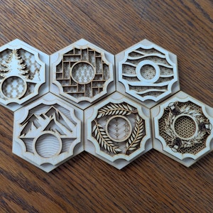 Puede incluir: Seis azulejos hexagonales de madera con intrincados diseños cortados con láser. Cada azulejo presenta un patrón diferente, que incluye árboles, un diseño geométrico, olas, montañas, trigo y una corona.