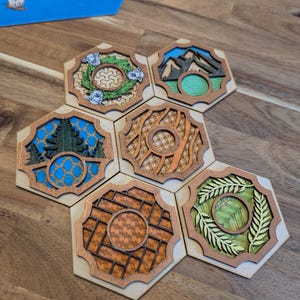 May include: Six hexagonal wooden tiles with different designs. Each tile features a different nature scene, including a wreath of flowers, mountains, a forest, a woven pattern, a grid pattern, and a wreath of leaves. The tiles are arranged in a circular pattern.