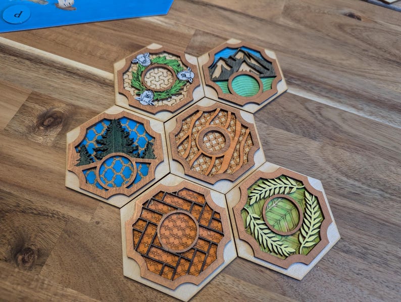 May include: Six hexagonal wooden tiles with different designs. Each tile features a different nature scene, including a wreath of flowers, mountains, a forest, a woven pattern, a grid pattern, and a wreath of leaves. The tiles are arranged in a circular pattern.