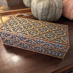 Peut inclure: Une boîte rectangulaire en bois présentant un motif en treillis complexe découpé au laser sur le couvercle et les côtés. Le revêtement en bois brun foncé contraste avec une doublure intérieure bleu clair visible à travers les découpes.