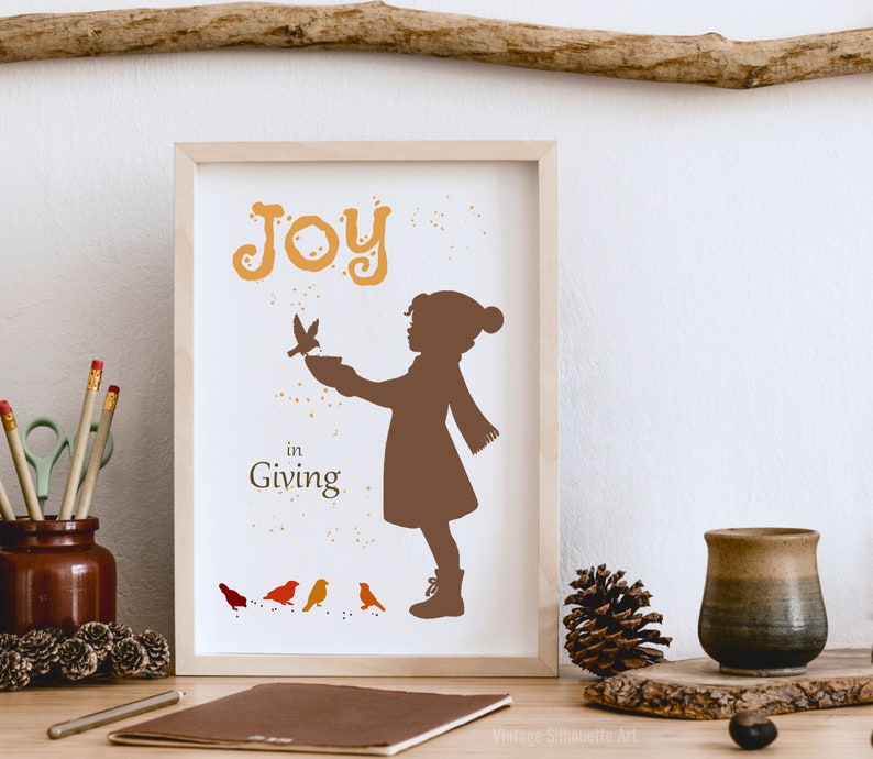 May include: A framed print with a brown silhouette of a child feeding birds. The print has the text "Joy in Giving" and is decorated with orange and yellow paint splatters.