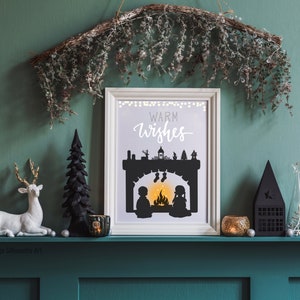 May include: A white framed print with a black silhouette of a fireplace with two figures standing in front of it. The text "Warm Wishes" is written in black cursive above the fireplace. The print is sitting on a green shelf with a variety of decorative items.
