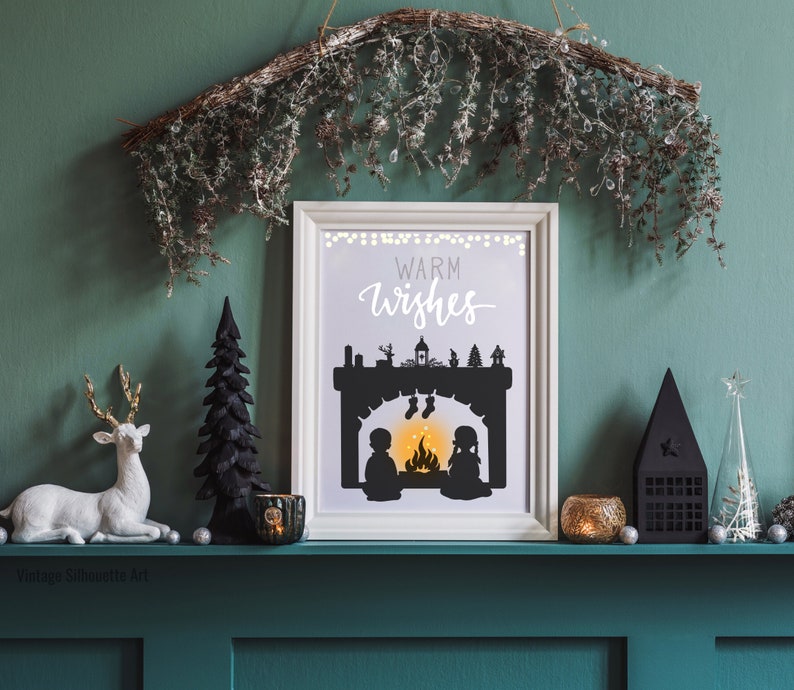 May include: A white framed print with a black silhouette of a fireplace with two figures standing in front of it. The text "Warm Wishes" is written in black cursive above the fireplace. The print is sitting on a green shelf with a variety of decorative items.