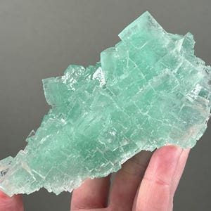 May include: A cluster of translucent, light green fluorite crystals featuring a cubic structure. The mineral specimen displays a vibrant mint-green hue with varying levels of clarity and sharp, geometric edges throughout the entire formation.