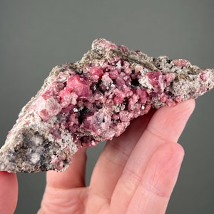 May include: A natural mineral specimen with bright pink rhodochrosite crystals on a grey and brown matrix. The crystals have a granular texture.