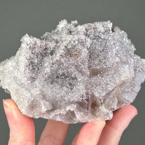 May include: A cluster of light purple and gray fluorite crystals. The crystals have a sparkling, textured surface and are held in a hand against a gray background. The mineral specimen has a natural, irregular shape.