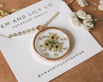 Könnte beinhalten: Eine goldene Kettenkette mit einem runden Anhänger, der gepresste weiße Blumen in klarem Harz enthält. Der Anhänger befindet sich auf einer weißen Karte mit dem Text "EM AND LILY CO handmade jewelry" und "@emandlilycollective".